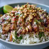 Sliced grilled chicken and fresh veggies rest on fluffy coconut rice, garnished with chopped peanuts and lime for a zesty finish.