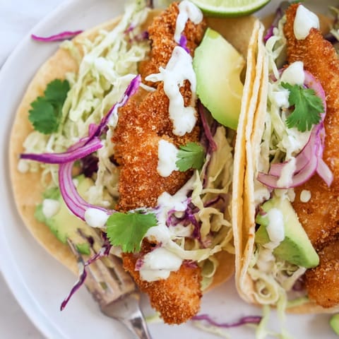 Perfectly fried Baja Fish Tacos garnished with avocado slices, cilantro, and red onion, paired with lime wedges on a rustic wooden table.