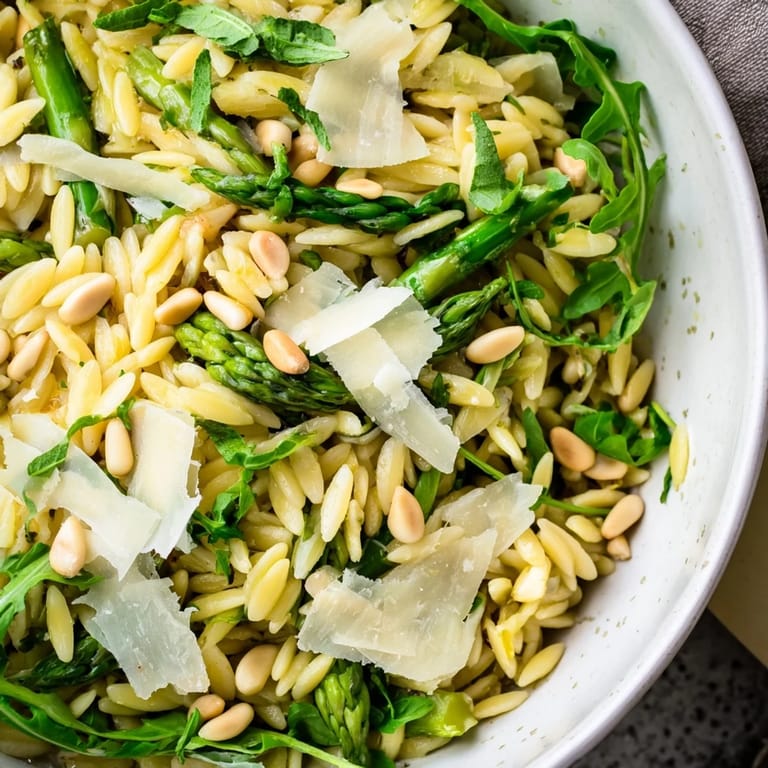 A close-up of Asparagus Lemon Orzo Salad shows glistening pasta, shaved asparagus, and shaved Parmesan, served in a white bowl with a lemon wedge.