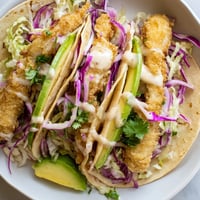 Golden-crisp battered fish fillets piled high on warm corn tortillas, topped with fresh green cabbage and creamy lime drizzle, ready to serve.